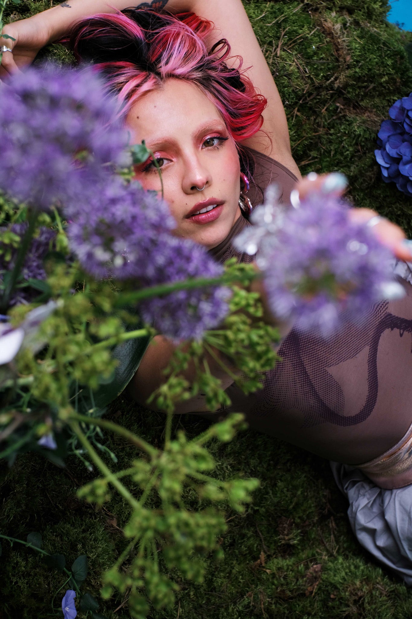 Natural skincare campaign photo — a person lying on moss, framed by purple wildflowers in a lush outdoor setting. QUINTA clean beauty, botanical and skin-first.