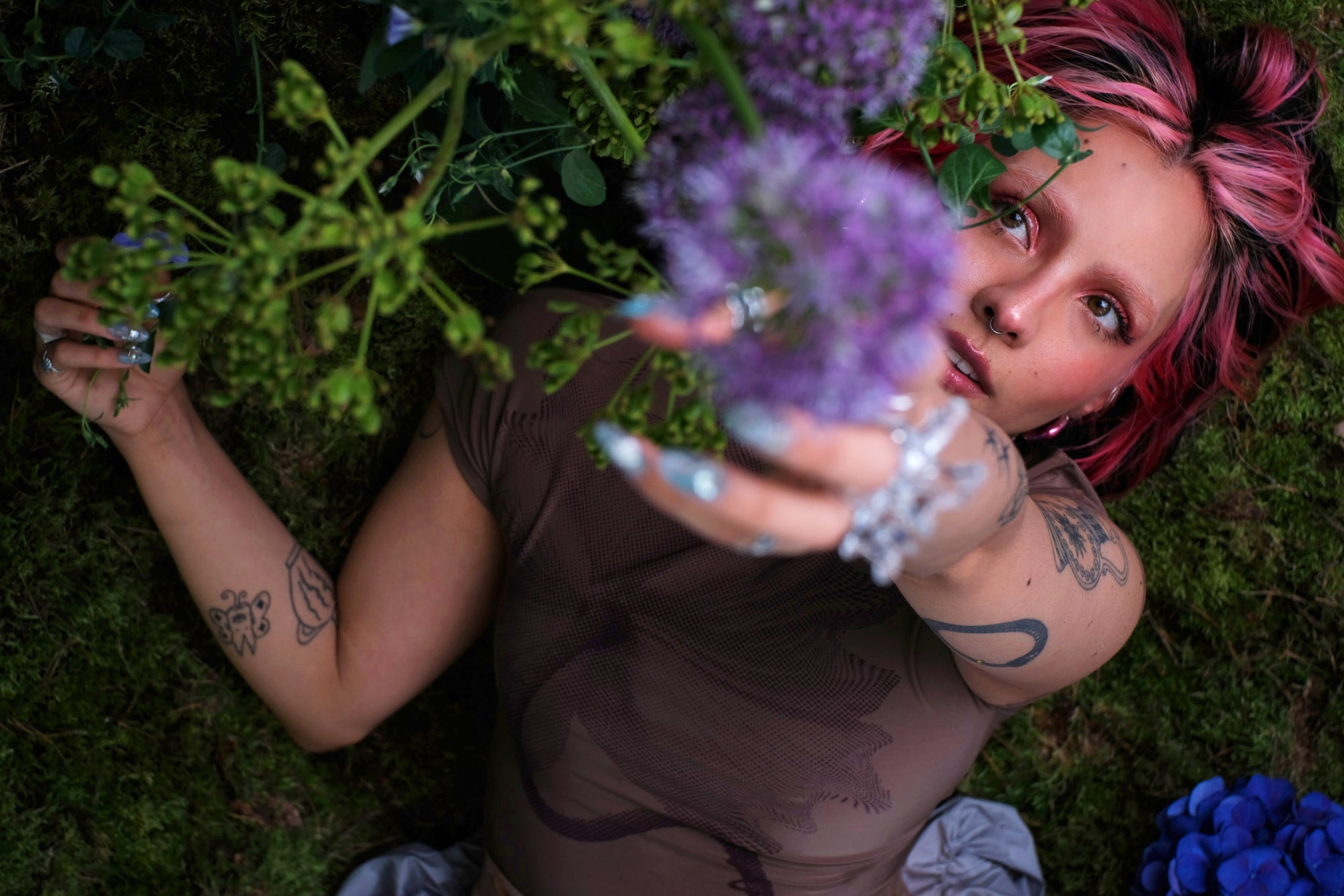 Person with colorful hair laying on green moss and reaching for a purple flower with one hand.
