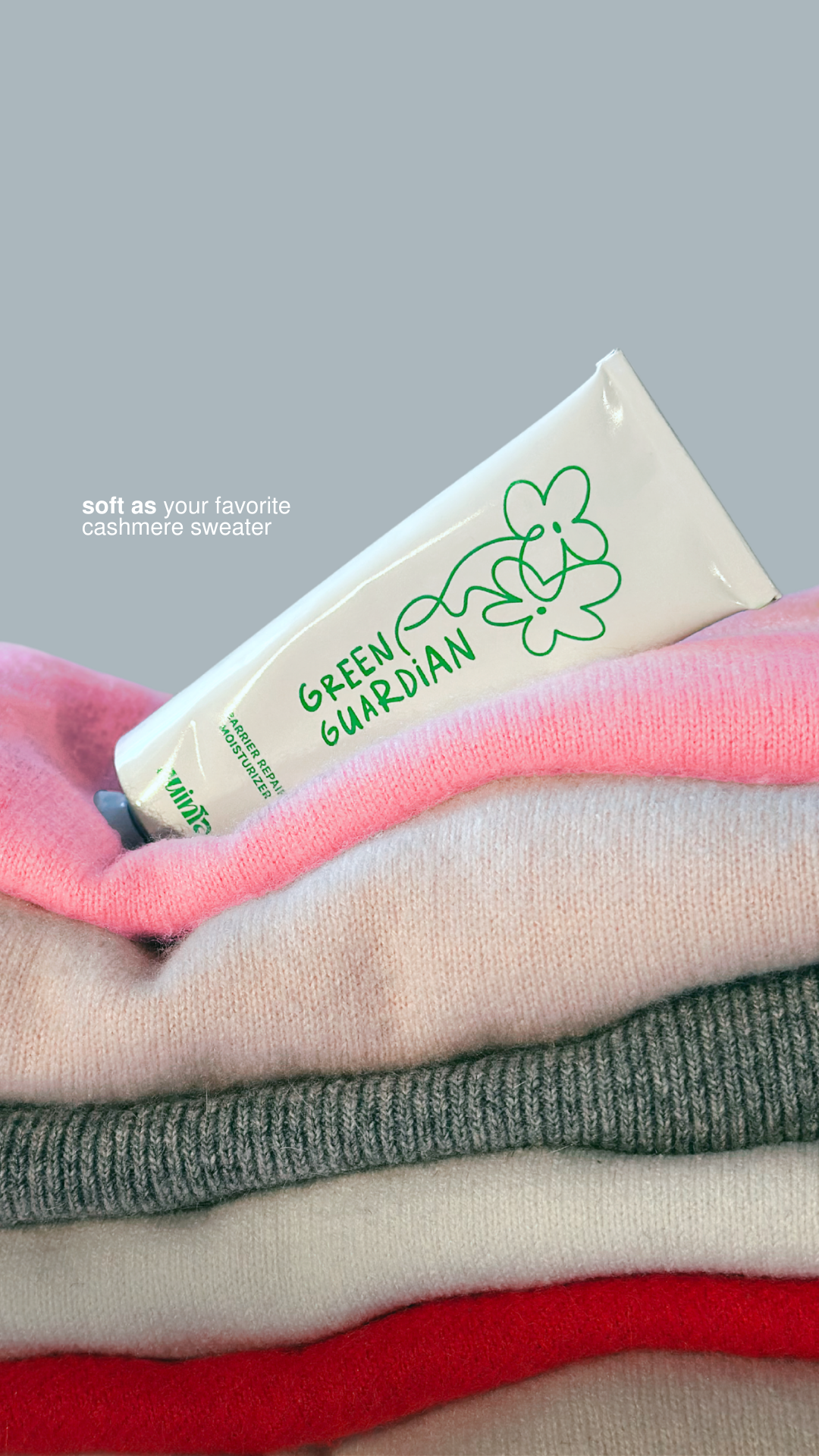 Tube of 'Green Guardian' product on a stack of colorful cashmere sweaters with a gray background.