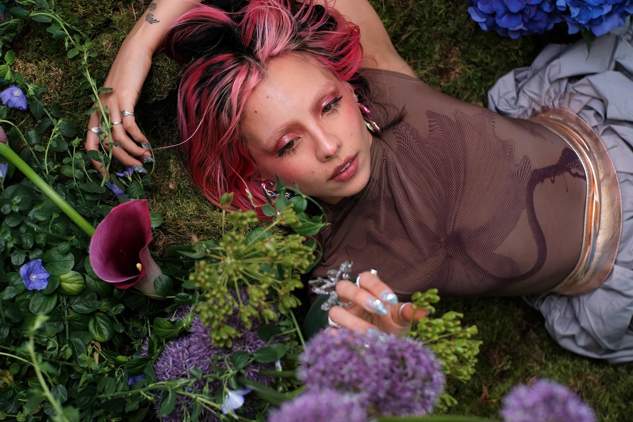 Natural skincare campaign photo — a person lying on moss, framed by purple wildflowers in a lush outdoor setting. QUINTA clean beauty, botanical and skin-first.