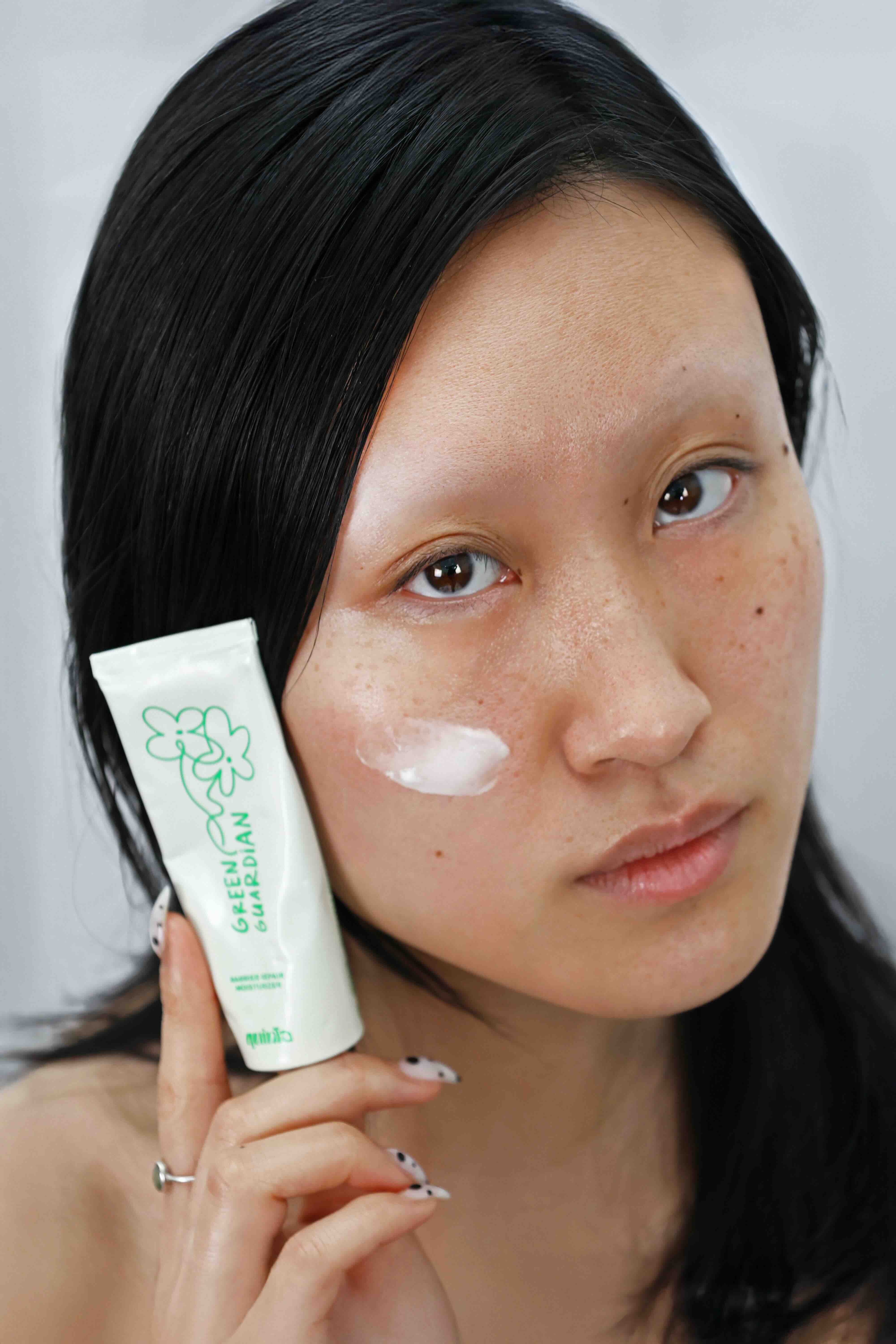 model with freckles holding quinta green guardian barrier repair moisturizer with cream applied to cheek